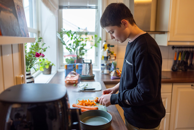 Tenåring står og ser ned, han er på et kjøkken og står med kjøkkenbenken hvor han holder på å skrelle gulrøtter.